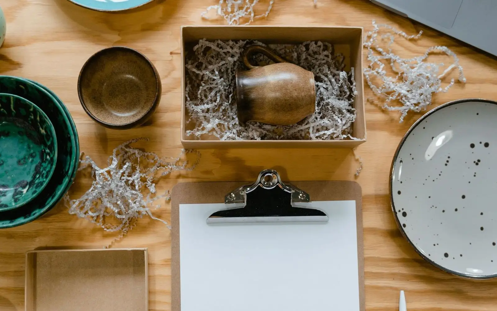 Ceramic bowls and plates beside a packing box of shredded paper and a clipboard, seen from above